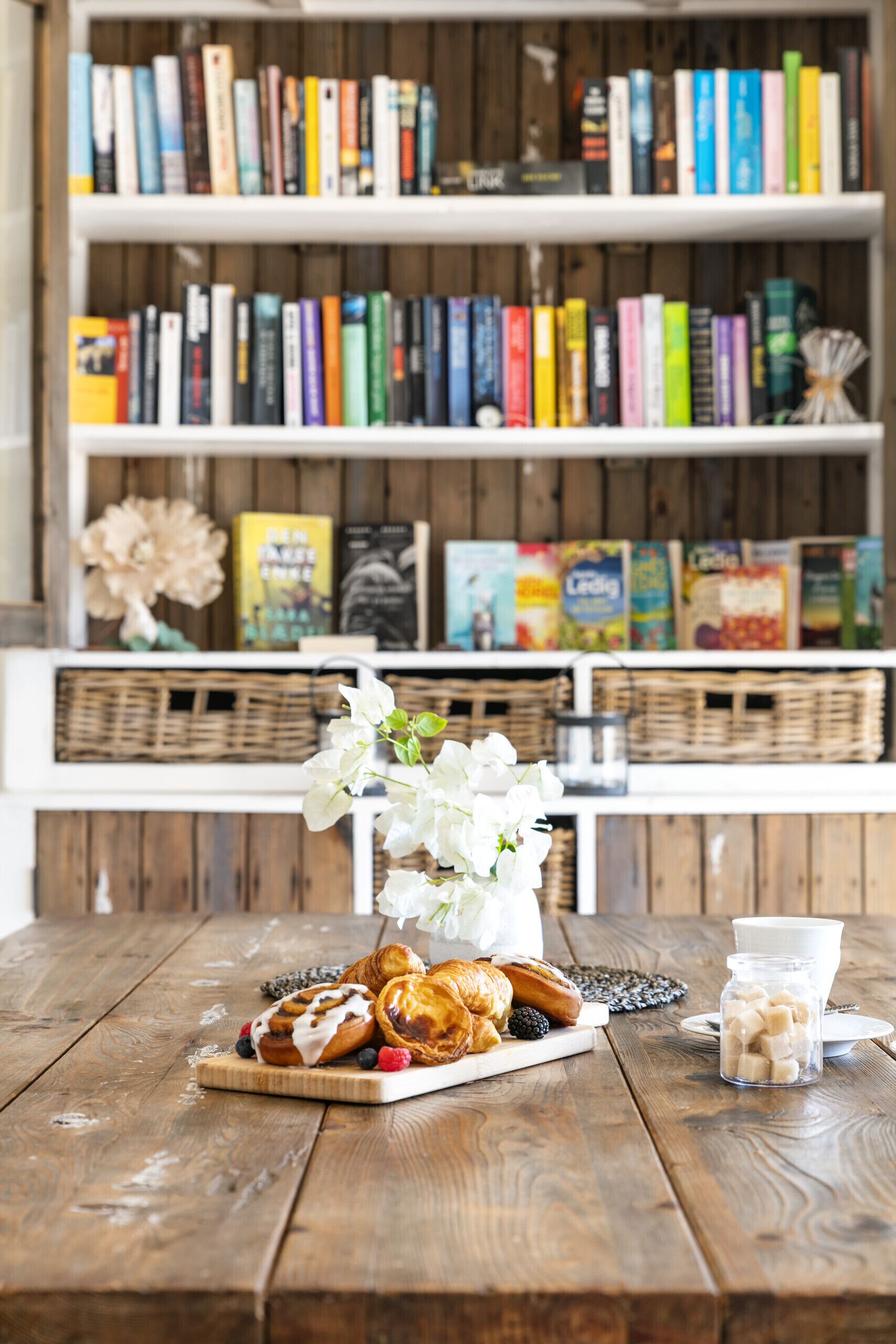 A wooden table with pastries, a vase of flowers, and a glass of milk. Behind the table, there is a bookshelf with various books and a wicker basket.