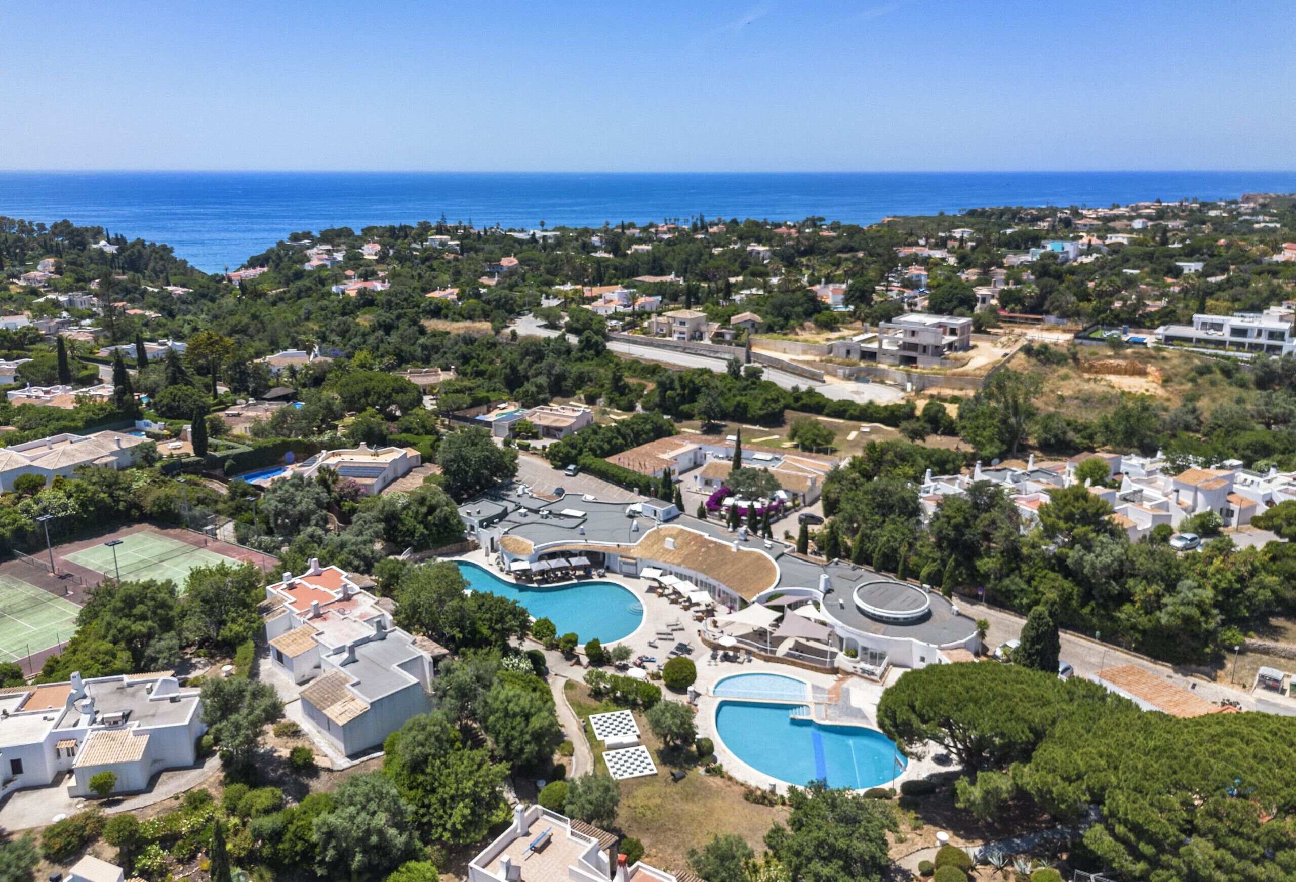 Vue aérienne d’un quartier résidentiel situé sur la côte, avec deux piscines, un court de tennis et différents types de maisons et d’immeubles.