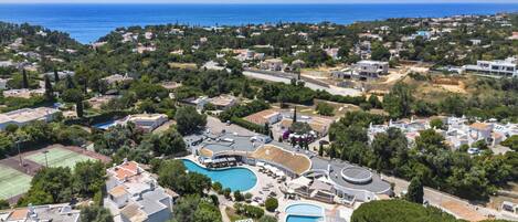 An aerial view of a coastal residential area with two swimming pools, a tennis court, and a variety of houses and buildings.