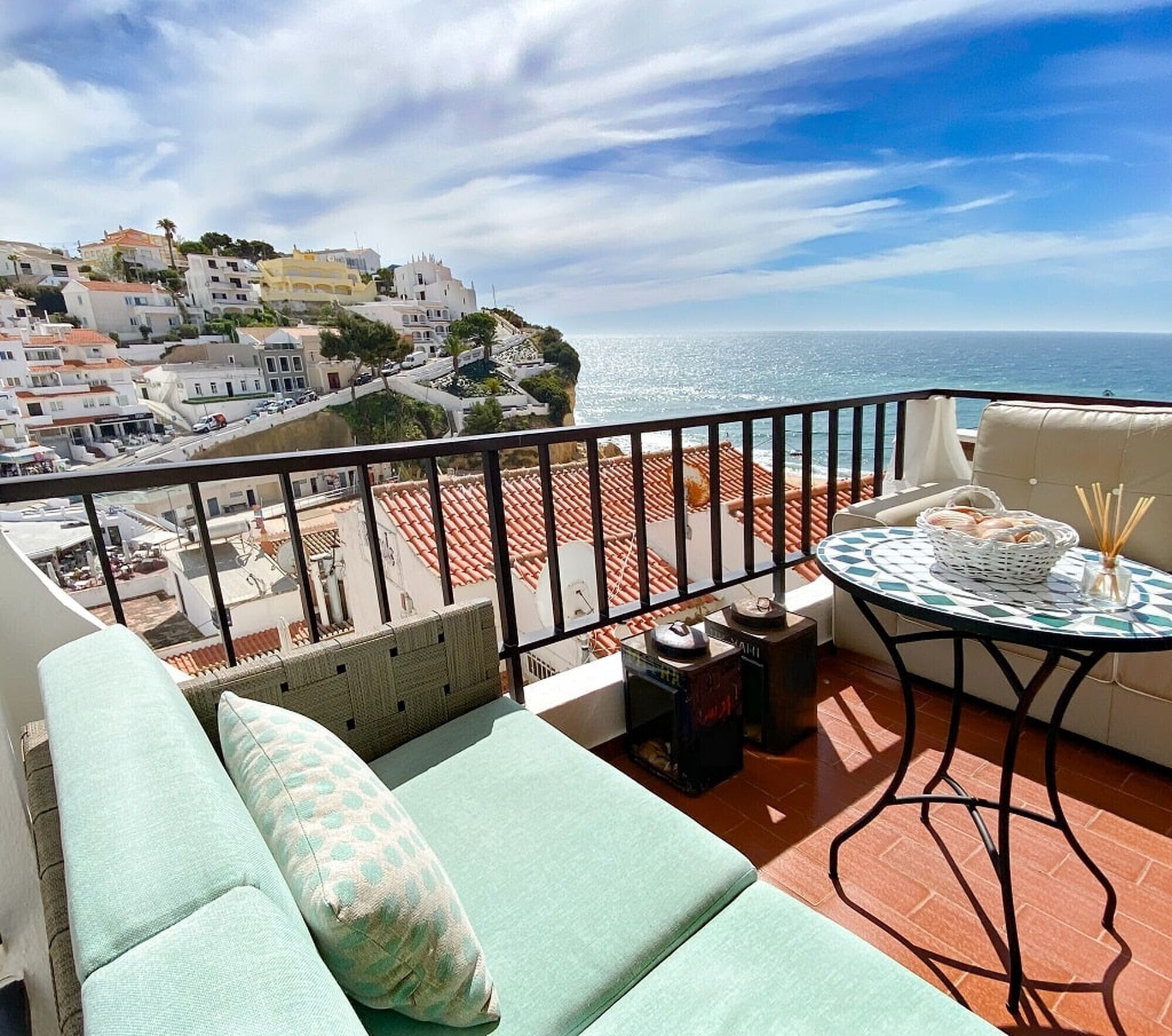 A balcony with a view of the sea and a coastal town. There is a sofa with cushions and a small table with a basket and a candle.