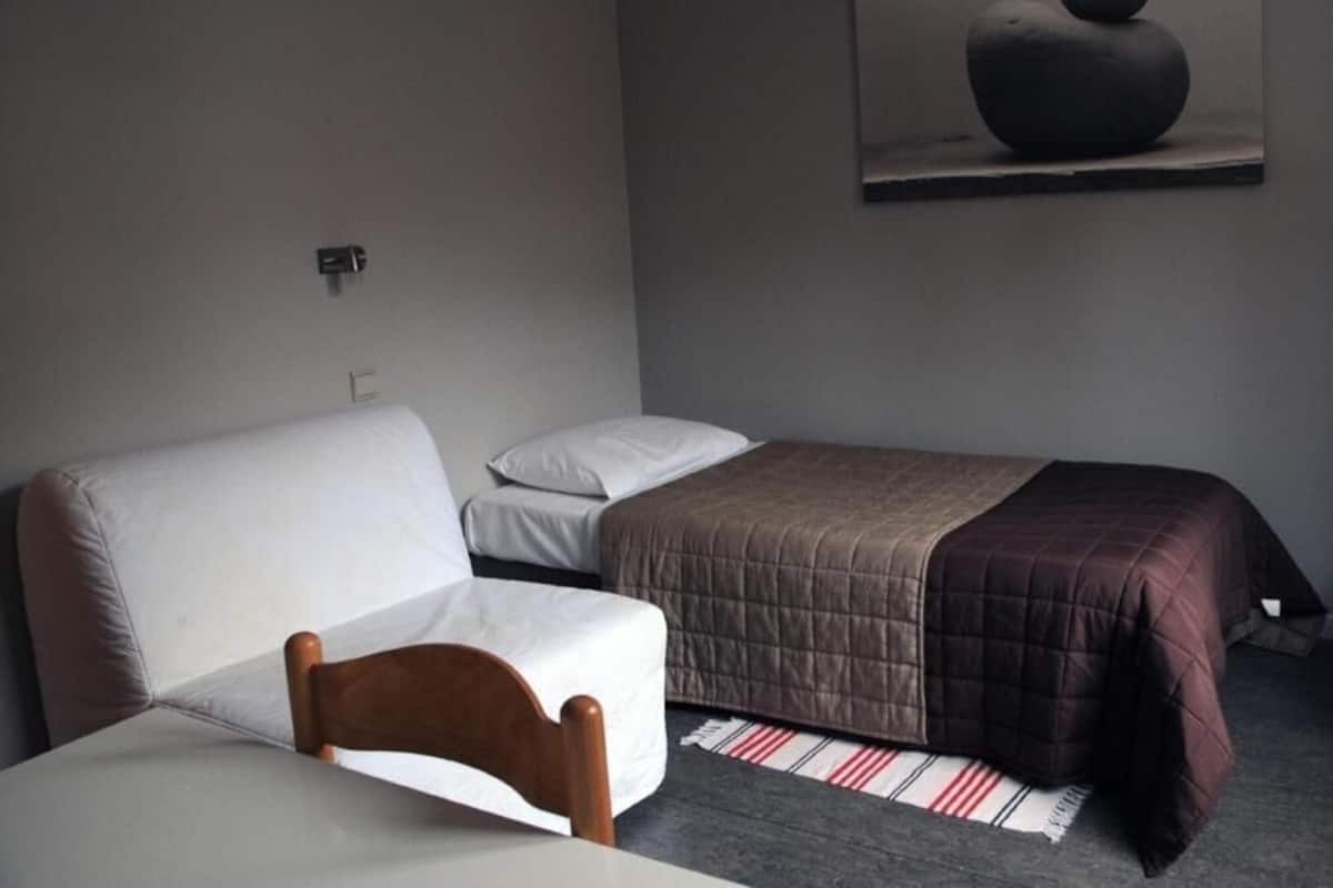 A bedroom with a bed, a chair, a bedside table, a wall-mounted artwork, and a power outlet.