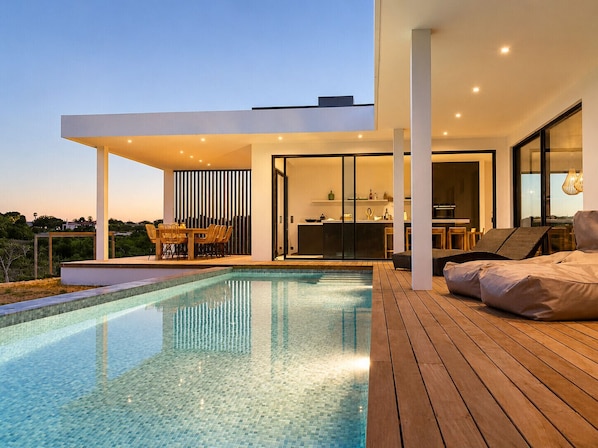 A modern outdoor pool area with a wooden deck, a dining area with chairs, and a kitchen visible through a glass door.