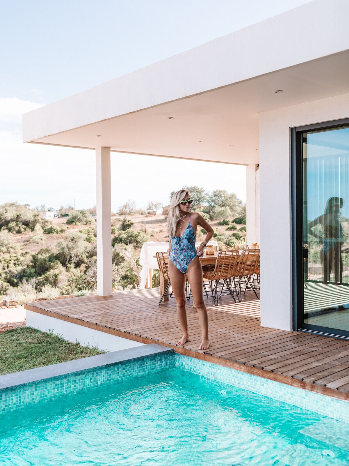 Un área de piscina exterior moderna con una terraza de madera, una mesa de comedor con sillas y vistas del paisaje circundante.
