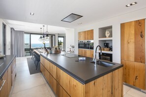 A modern kitchen with a large island, wooden cabinets, and a view of the ocean. - Villa da Raposa - Stunning ocean view villa! (Lagos)