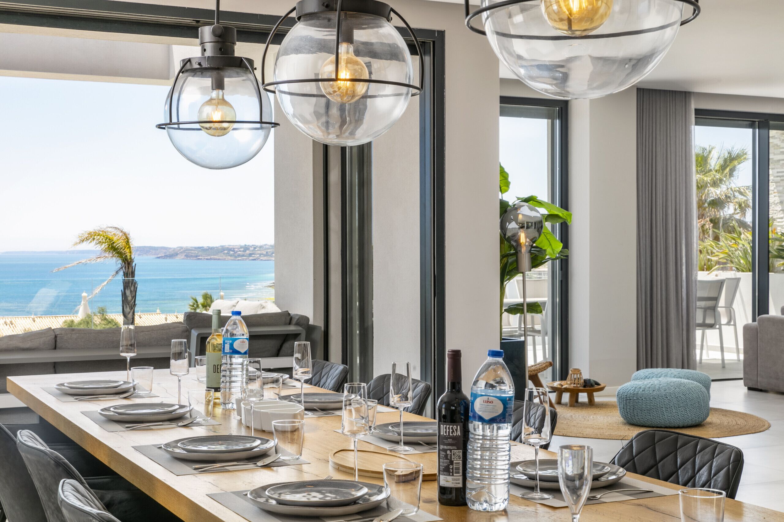 A dining area with a view of the ocean, a long table set for a meal, and modern pendant lighting.