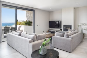 A modern living room with two grey sofas, a flat-screen TV, a fireplace, and a large sliding glass door leading to a balcony with a view of the sea. - Villa da Raposa - Stunning ocean view villa! (Lagos)