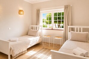 A room with two beds, a window with curtains, a small table, and a wall-mounted light fixture.