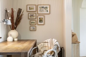 Ein Zimmer mit einem Holztisch, einem weißen Stuhl, einer Lampe, einer Vase mit Trockenblumen und mehreren gerahmten Bildern an der Wand.