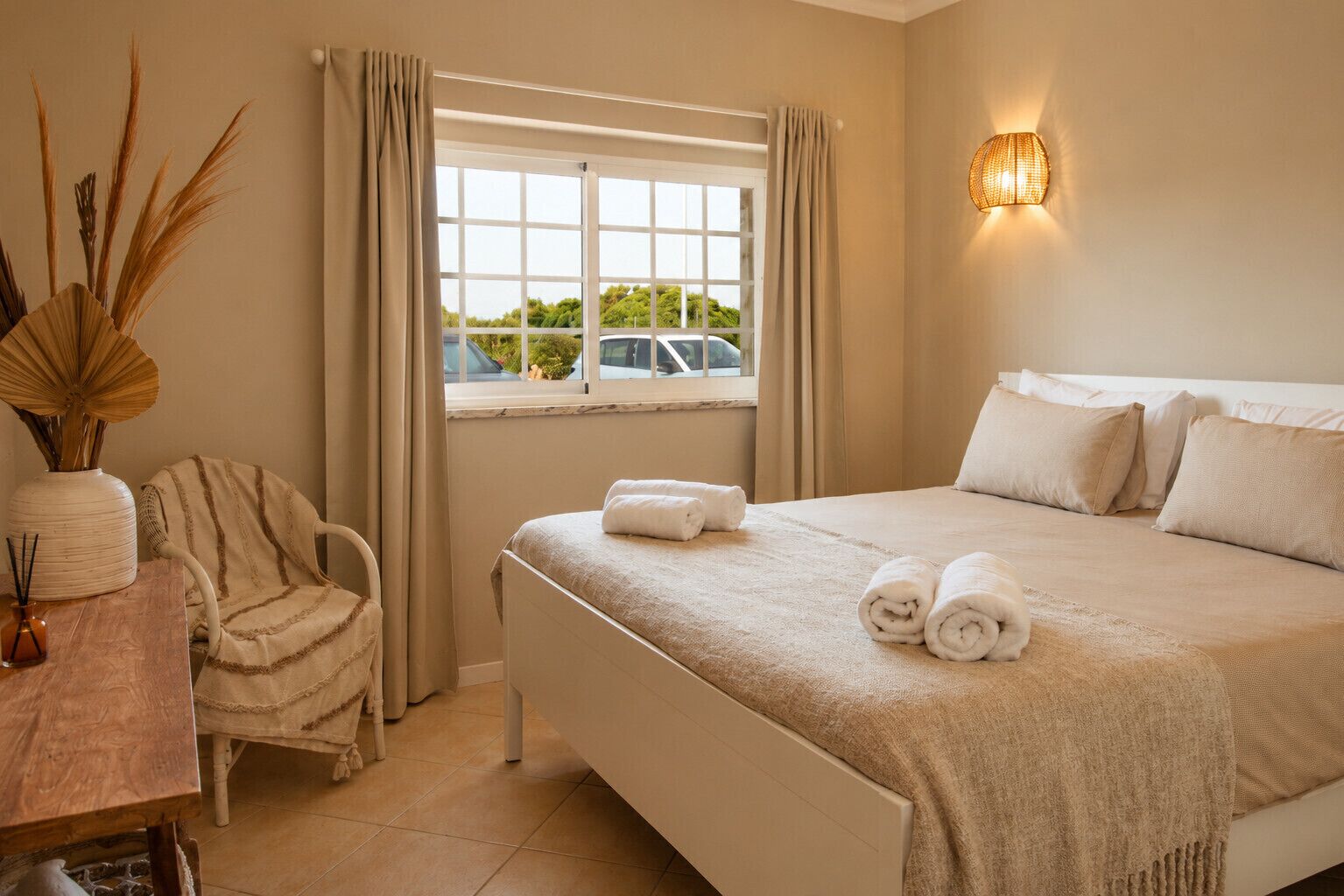 Une chambre d’hôtel avec un lit, une chaise, un bureau, un vase contenant des plantes séchées et une fenêtre avec des rideaux.