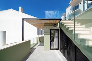 A narrow outdoor corridor with a glass door leading to a covered area, a staircase, and a white building in the background. - The Beach House Carvoeiro - Cliffside Apartment (Carvoeiro)