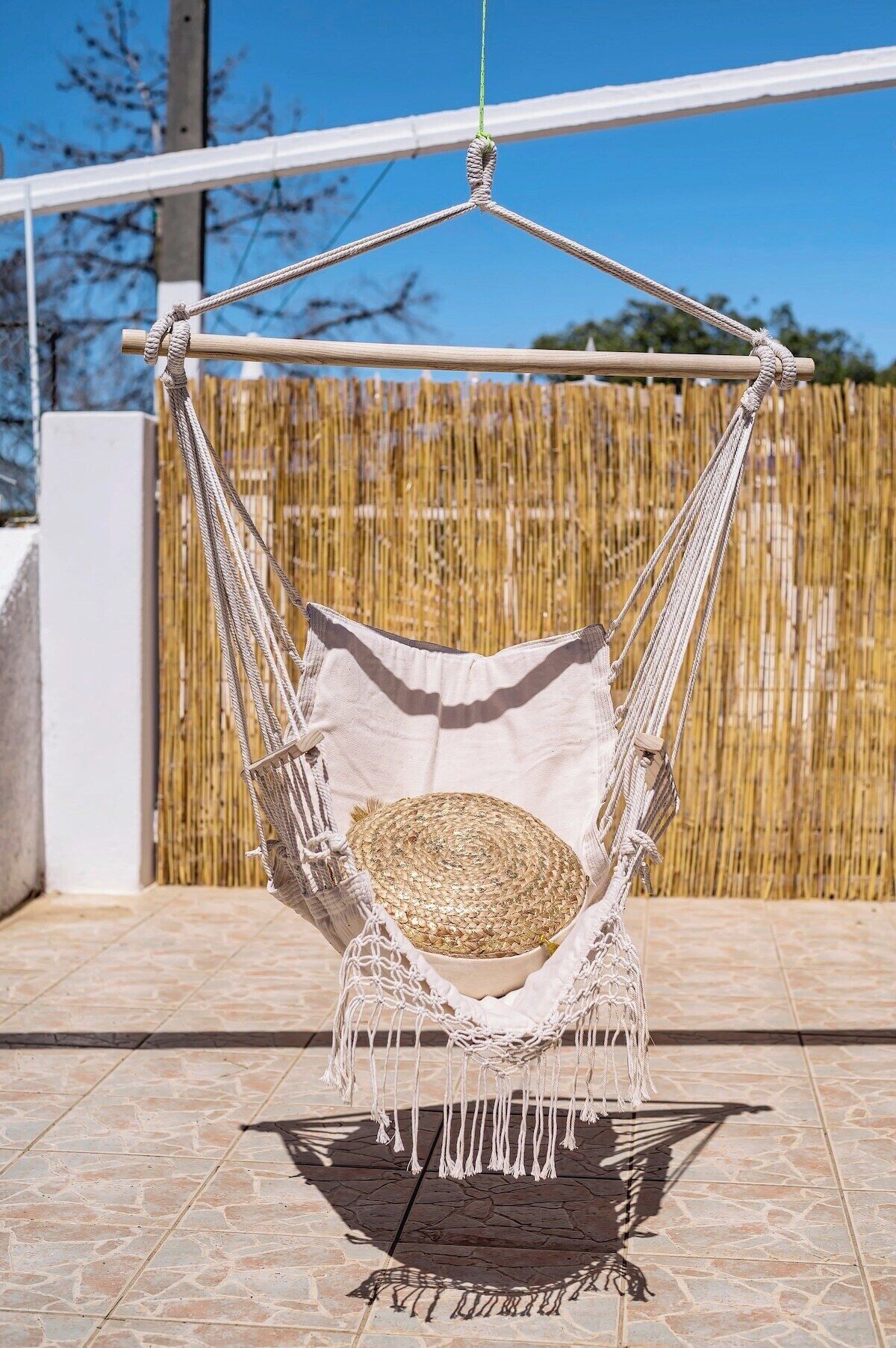 Un fauteuil suspendu en forme de hamac, orné de franges en macramé, un panier tressé et un coussin blanc, disposés sur une terrasse carrelée, avec une clôture en bambou en arrière-plan.