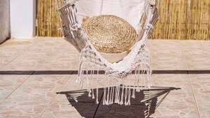 Ein Hängestuhl mit Fransenoptik, ein gewebter Korb und ein weißes Kissen auf einer gefliesten Terrasse mit einem Bambuszaun im Hintergrund.