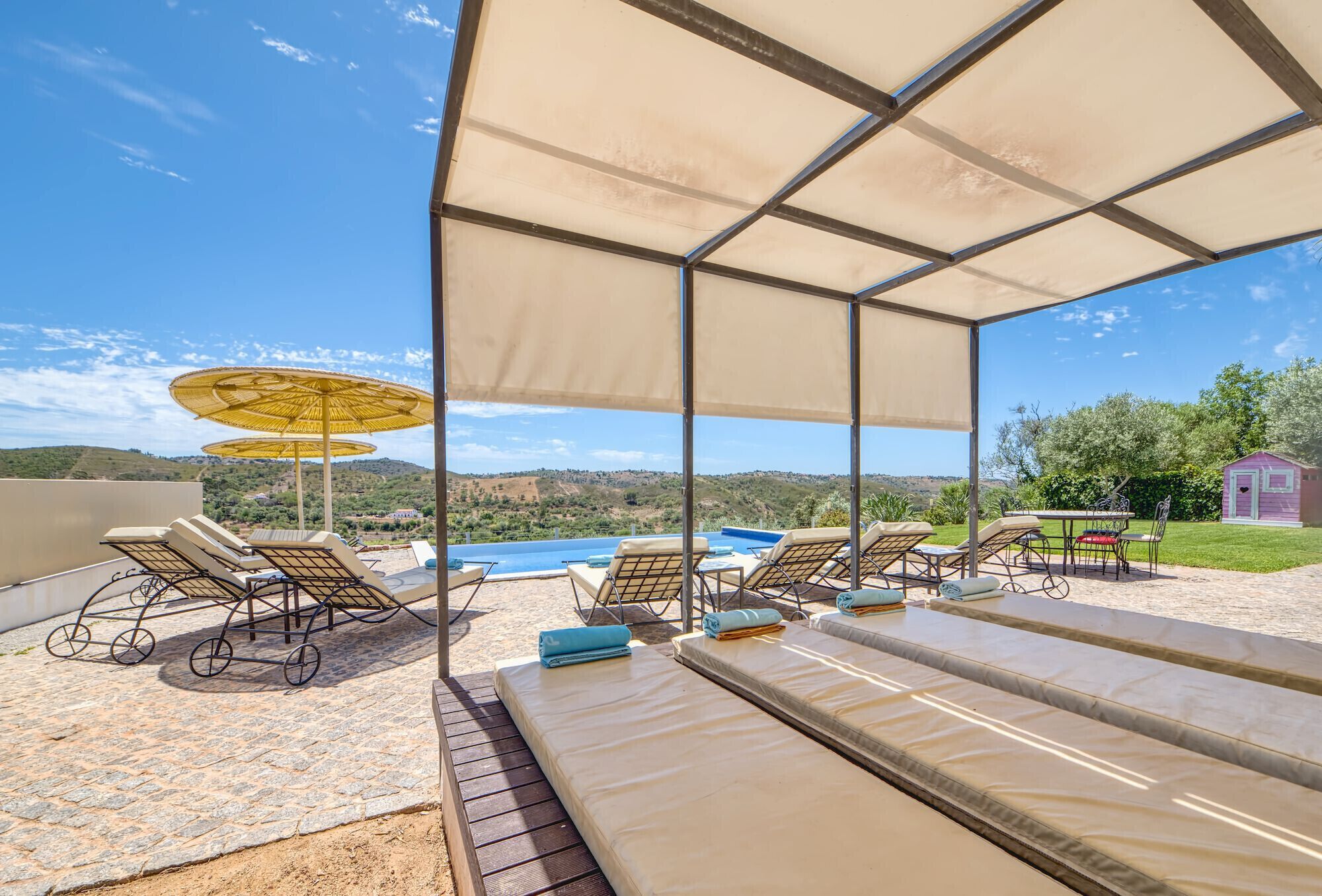 A sunlit outdoor lounge area with lounge chairs and umbrellas, overlooking a scenic landscape and a body of water.
