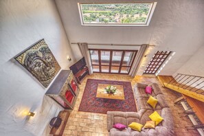 A spacious living area with a high ceiling, a large window offering a view of the countryside, a cozy sofa with cushions, a patterned rug, and a wooden coffee table. - Villa Fuzeiros - Amazing villa in a green valley! (São Bartolomeu de Messines)