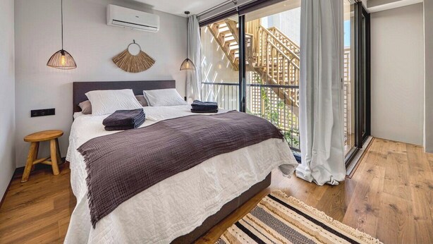 Une chambre moderne avec un grand lit, un petit tabouret, un luminaire suspendu et une vue sur un escalier en bois, visible à travers une porte vitrée.