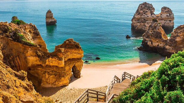 Un paisaje costero con formaciones rocosas, una playa arenosa y una escalera de madera que desciende hacia la orilla.