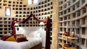 A bedroom with a stone wall, a bed with white linens, red curtains, and a metal shelf with books and a picture frame. - Studio Michel (Pichanges)