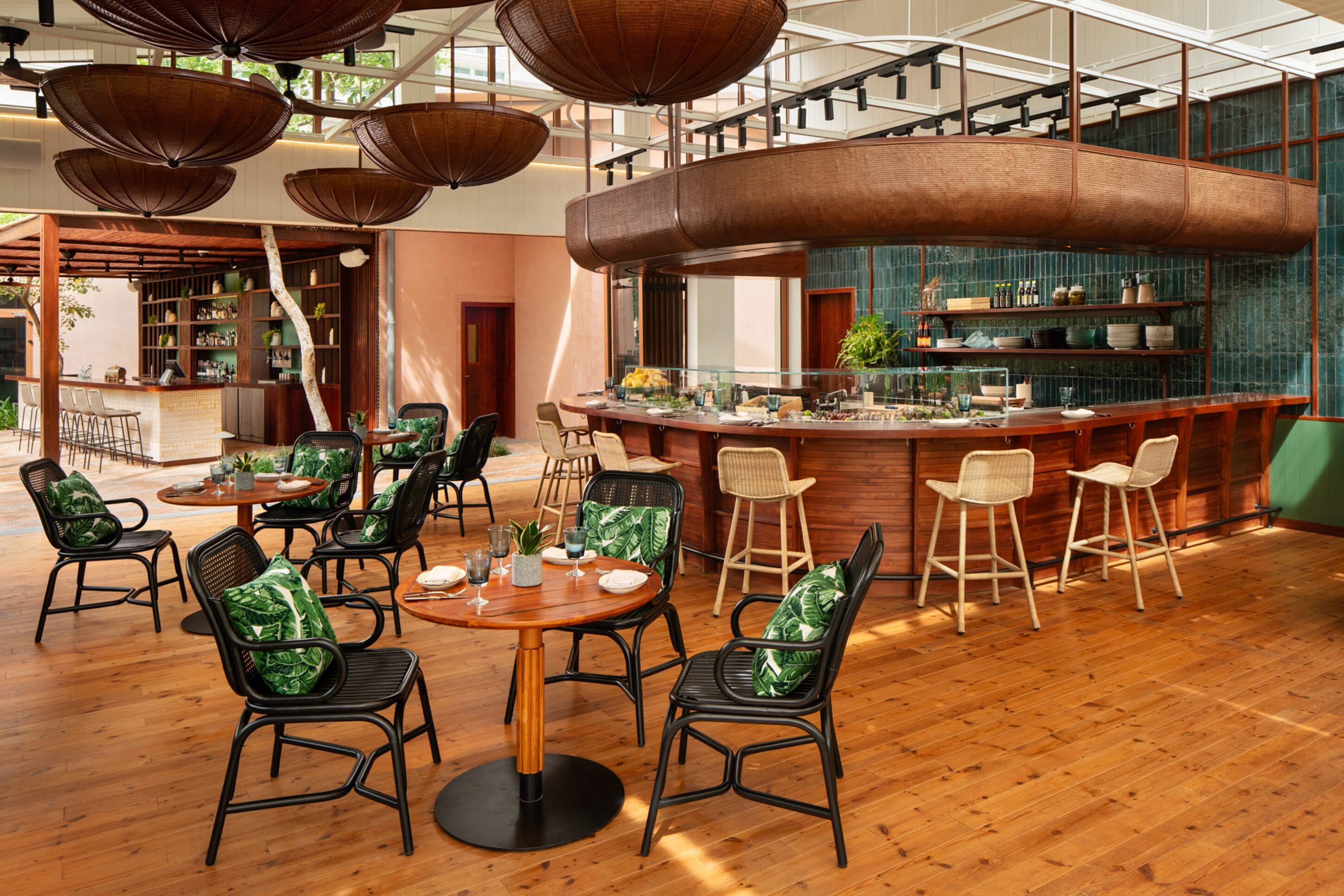 A modern restaurant with wooden floors, green cushioned chairs, and a curved bar area.