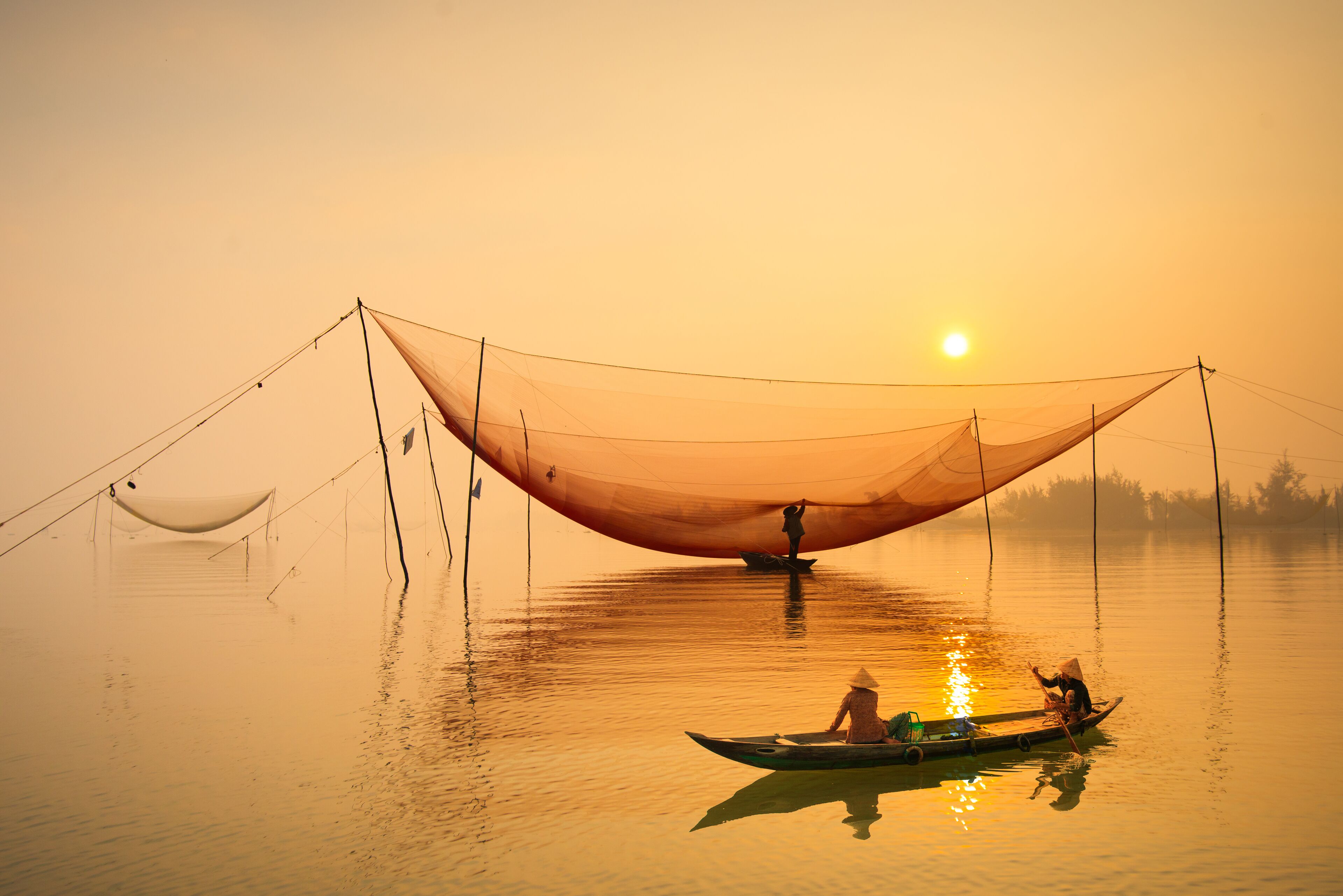 Um lago calmo ao pôr do sol, com uma grande estrutura de rede e um pequeno barco com pessoas.