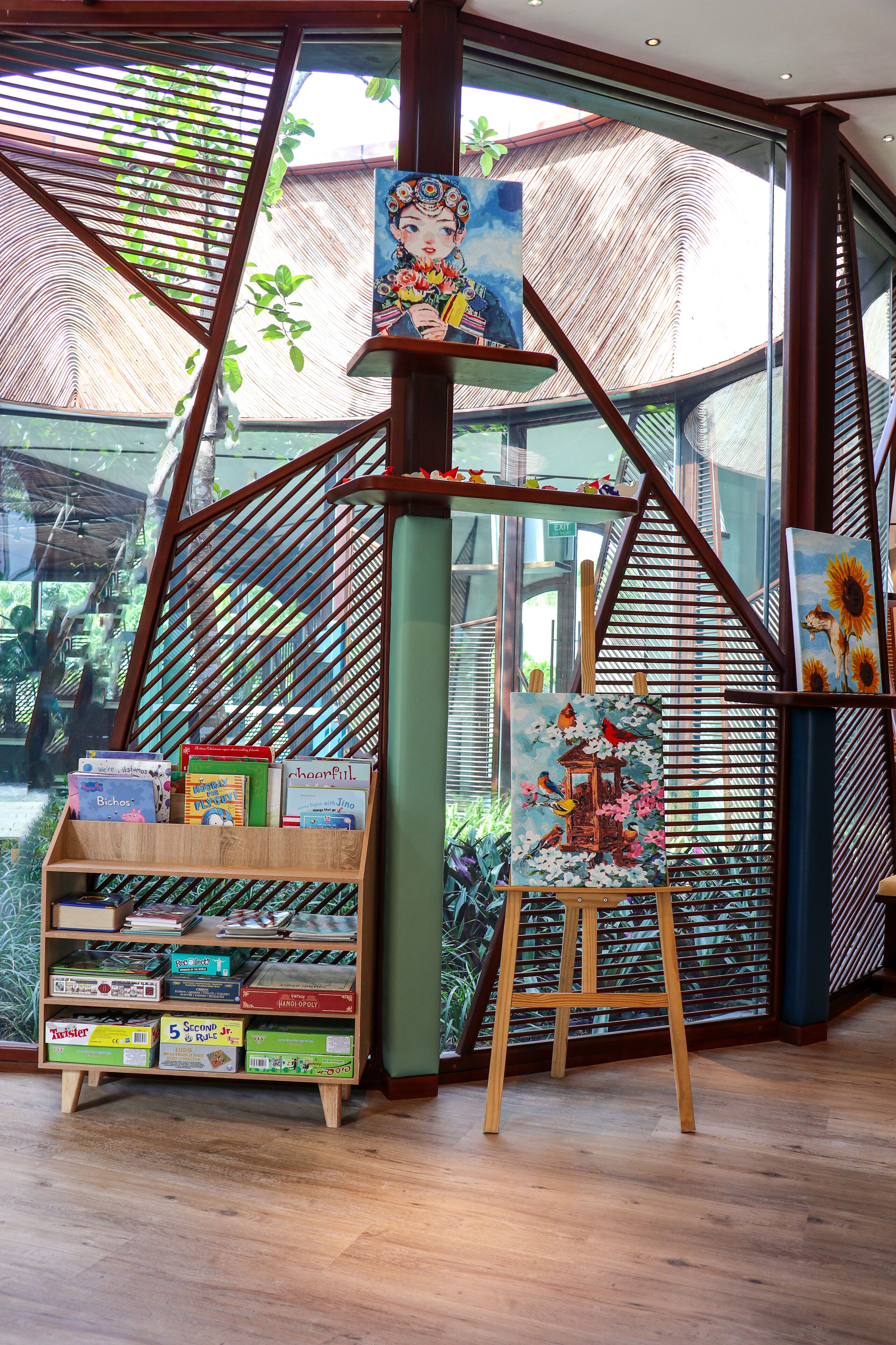 A room with a wooden bookshelf filled with books, a painting on an easel, and a large window with a view of greenery.