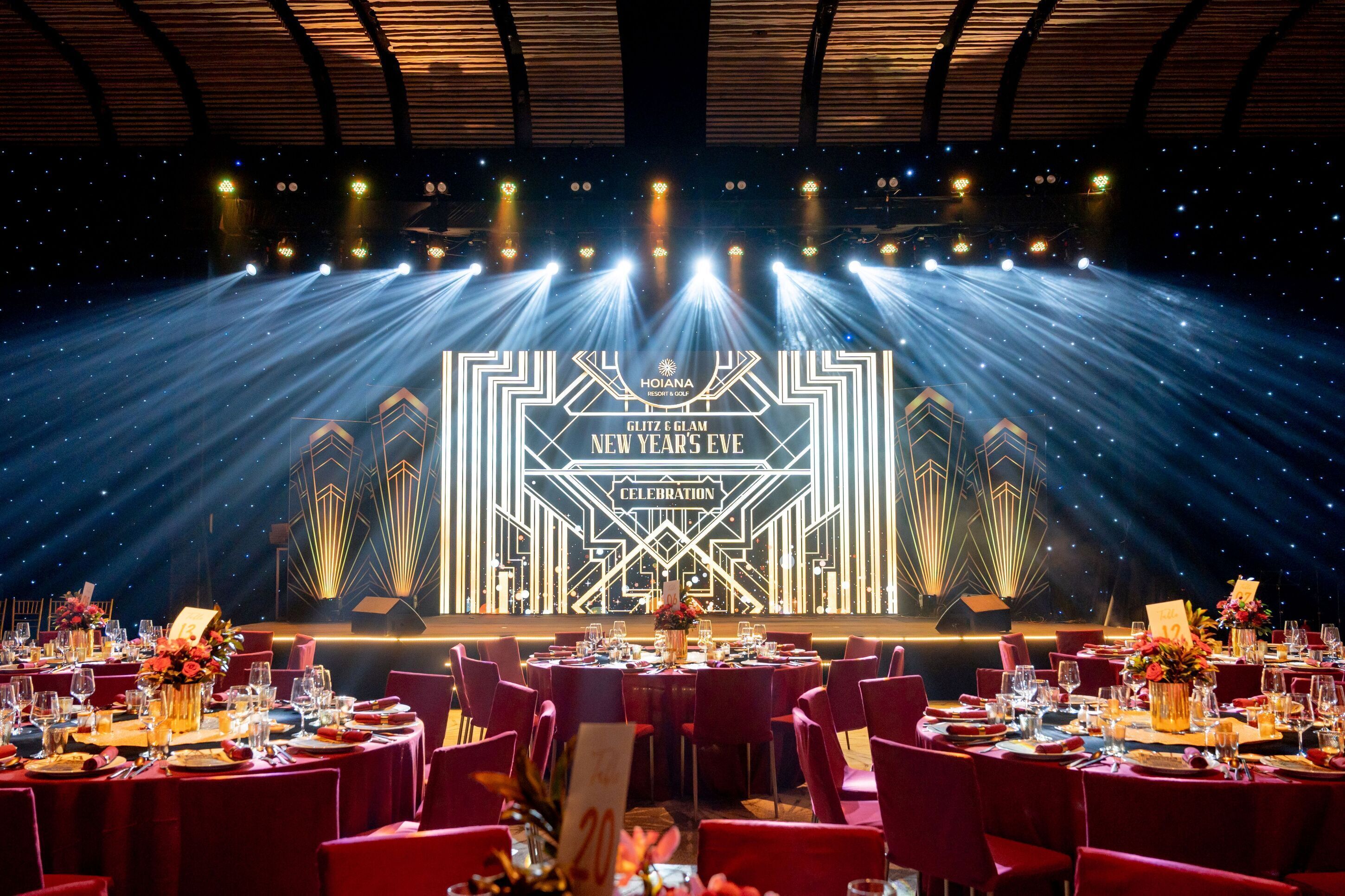 A formal event space with tables set for a celebration, featuring a stage with a geometric light design and the text "HOIANA GLITZ & GLAM NEW YEAR'S Eve CELEBRATION".