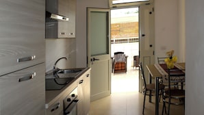 A modern kitchen with wooden cabinets, a stainless steel sink, and an oven. There is a dining area with a table and chairs, and a view of the outside through an open door. - Tenuta Paifer (Otranto)