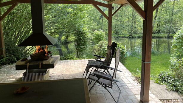 Une terrasse extérieure dotée d’un poêle à bois, d’un sol en pierre et offrant une vue sur un lac entouré d’arbres.