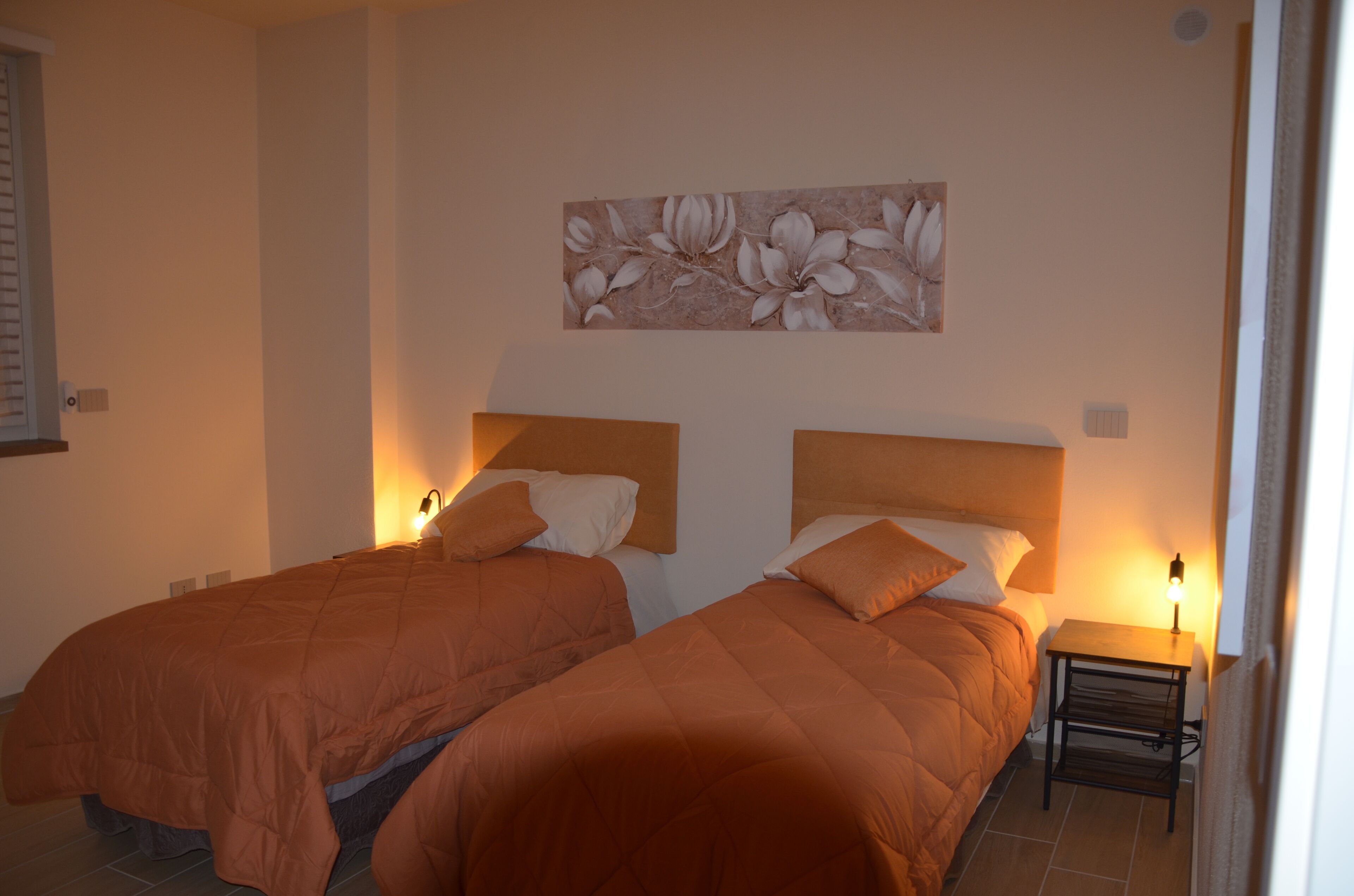 Una habitación con dos camas, cada una con cubrecama naranja y almohadas blancas. Hay una mesita de noche con una lámpara junto a una de las camas.