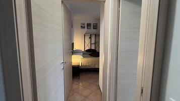 A narrow hallway leading to a bedroom with a bed, a nightstand, and a wall-mounted shelf.