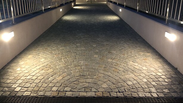 Uma passagem pavimentada, ladeada por grades de metal e iluminada por luzes embutidas na parede, conduz a uma entrada de edifício.