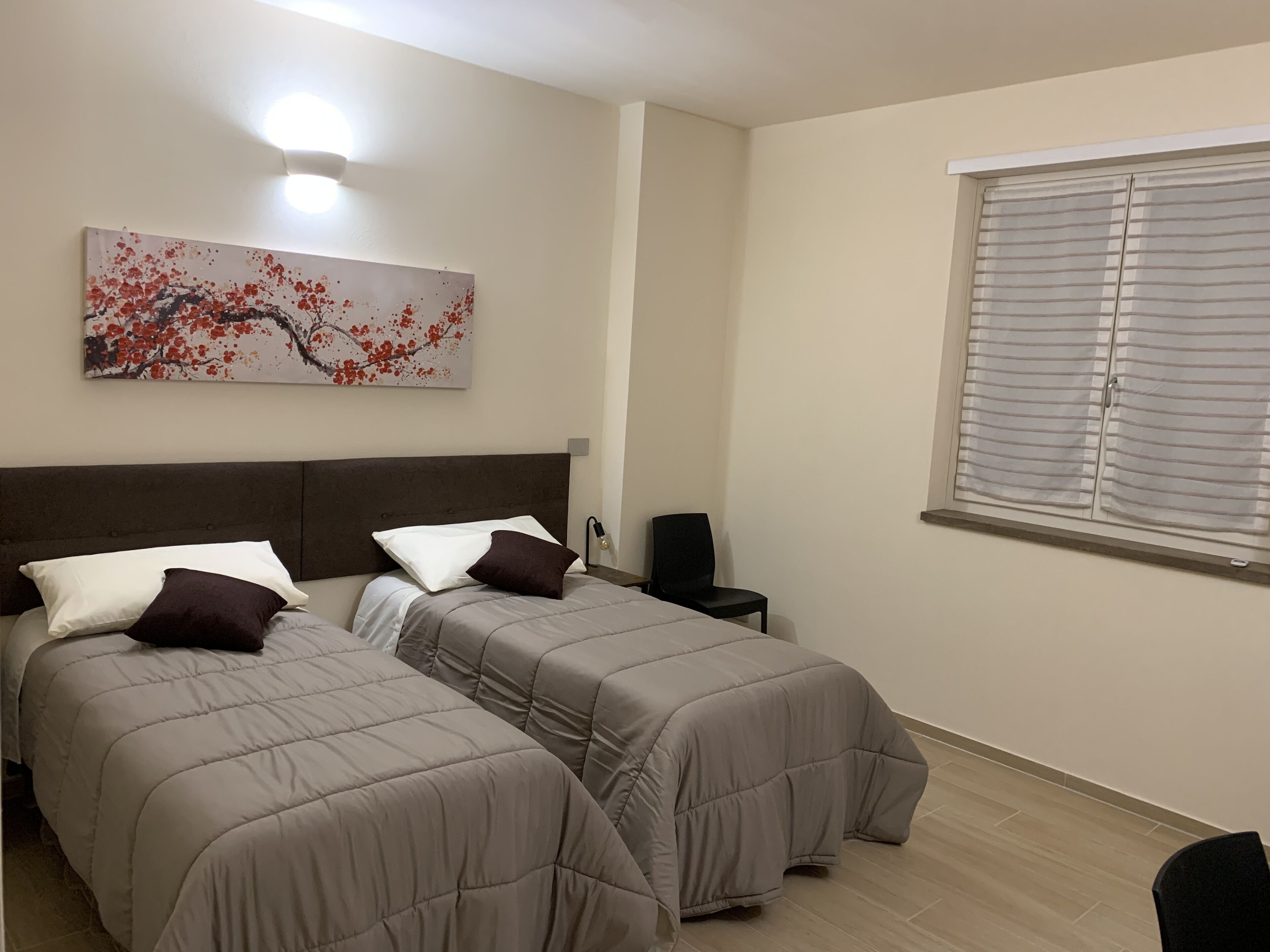 Una camera da letto con due letti, un comodino, una sedia, una finestra con persiane e una lampada a parete.
