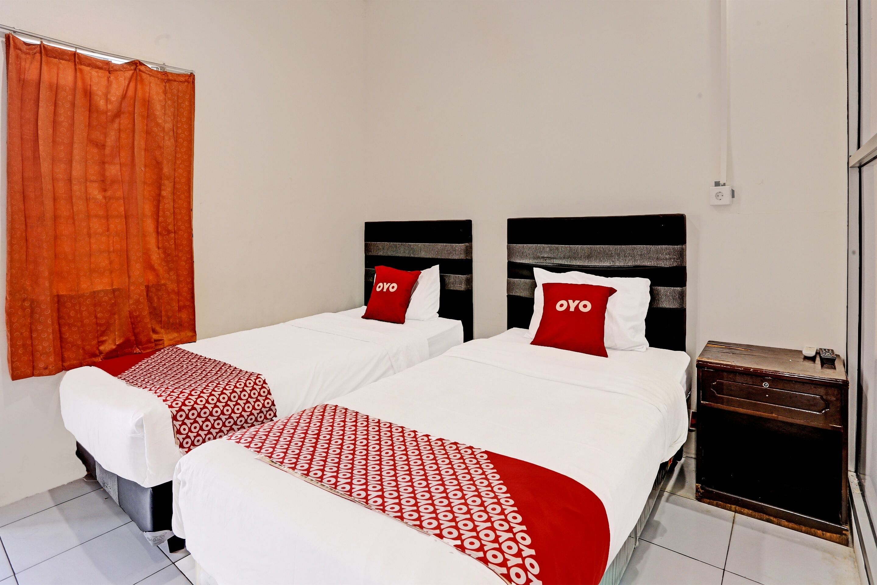 A hotel room with two beds, each with a red and white patterned bedspread, a wooden nightstand, and a red curtain.