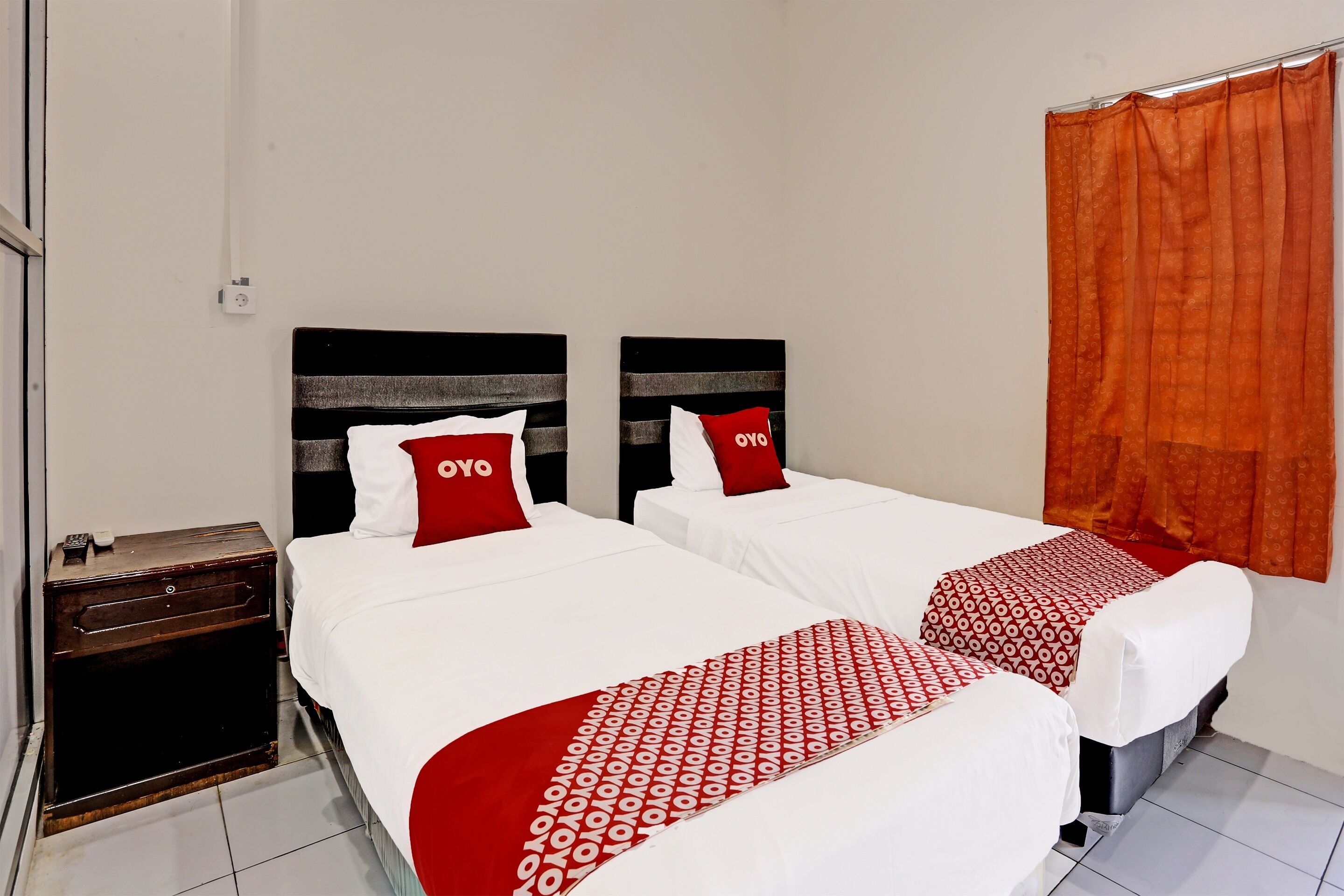 A hotel room with two beds, each with a white bedspread and red pillows. There is a wooden nightstand with a small table and a lamp.