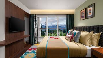 Habitación de hotel con cama, televisor, balcón con vistas a la montaña y ventana con cortinas.