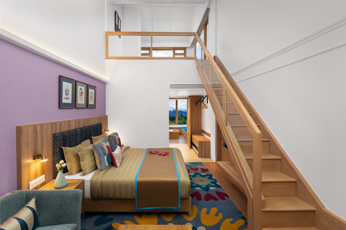 A modern hotel room with a bed, a wooden staircase, and a view of the outdoors.