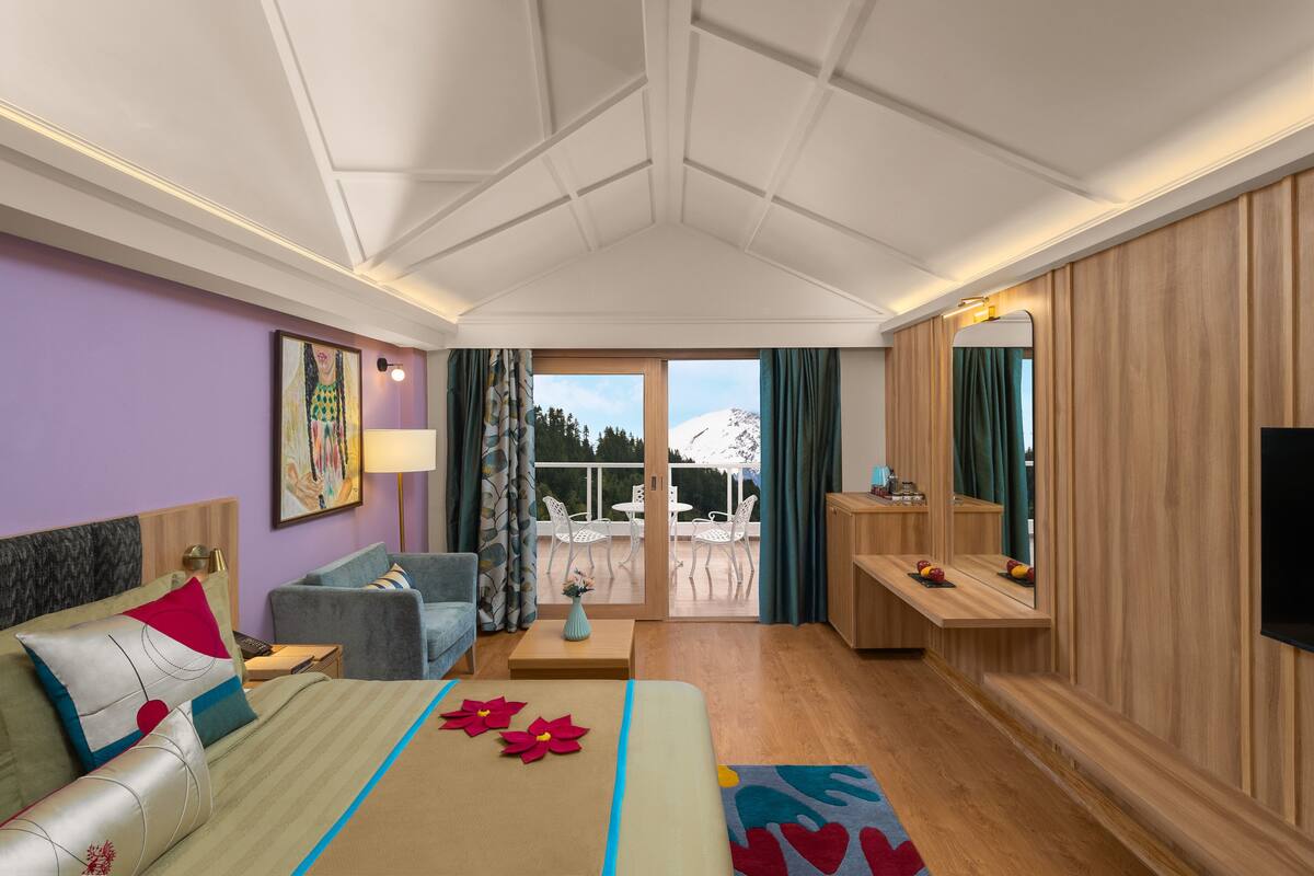 A hotel room with a bed, a sofa, a small table, a TV, and a balcony with a view of the mountains.