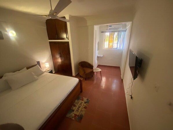 Un dormitori amb llit, ventilador al sostre, armari, taula petita amb cadira, finestra amb cortines i porta que condueix a una altra estança.