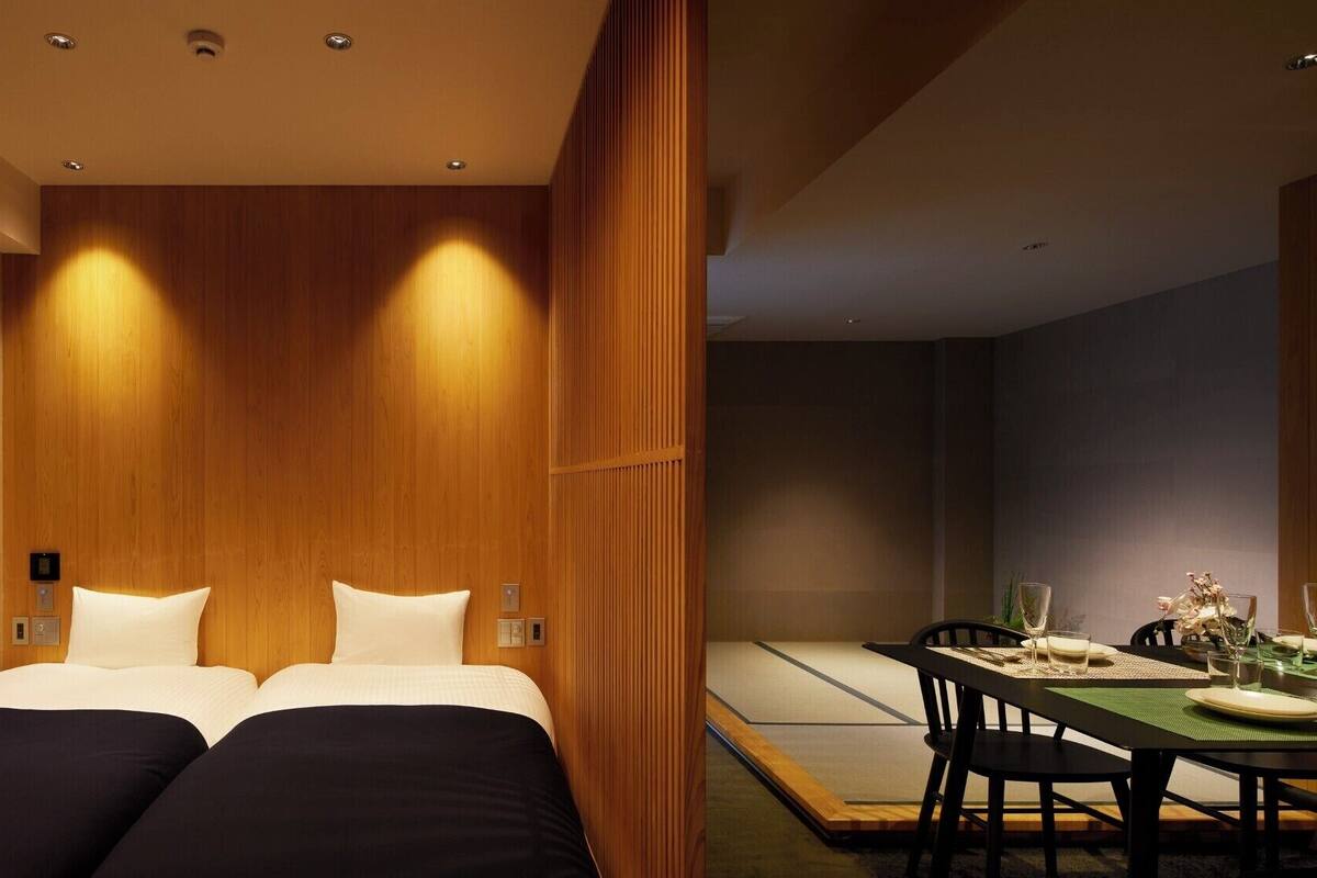 Een moderne hotelkamer met een houten accentmuur, een bed met witte lakens, een eethoek met een tafel gedekt voor twee personen, en een klein bijzettafeltje met een bloevas.