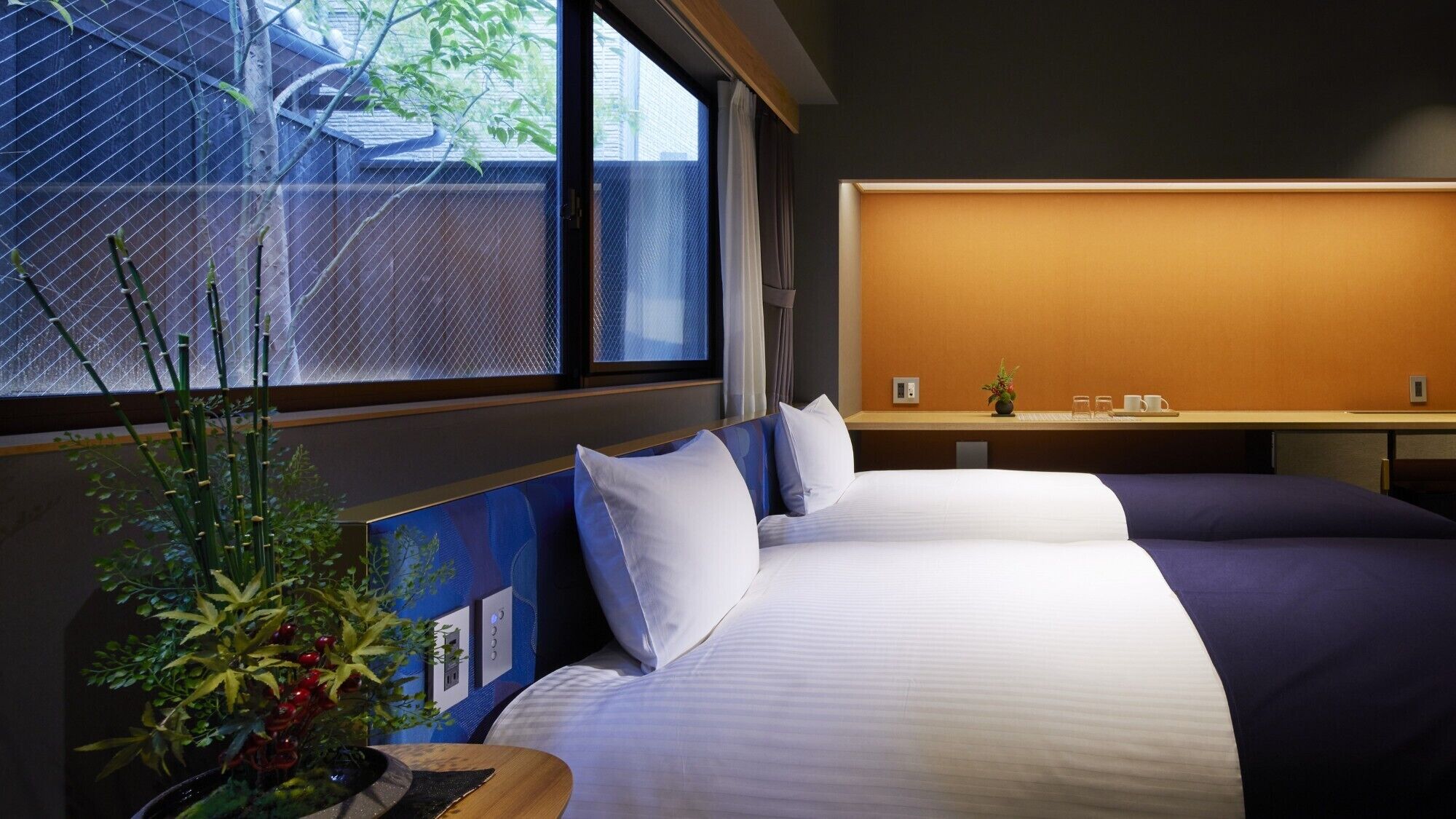 A hotel room with two beds, a window with a view of greenery, a small table with a plant, and a wall-mounted shelf with a vase and cups.