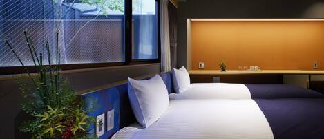 A hotel room with two beds, a window with a view of greenery, a small table with a plant, and a wall-mounted shelf with a vase and cups.