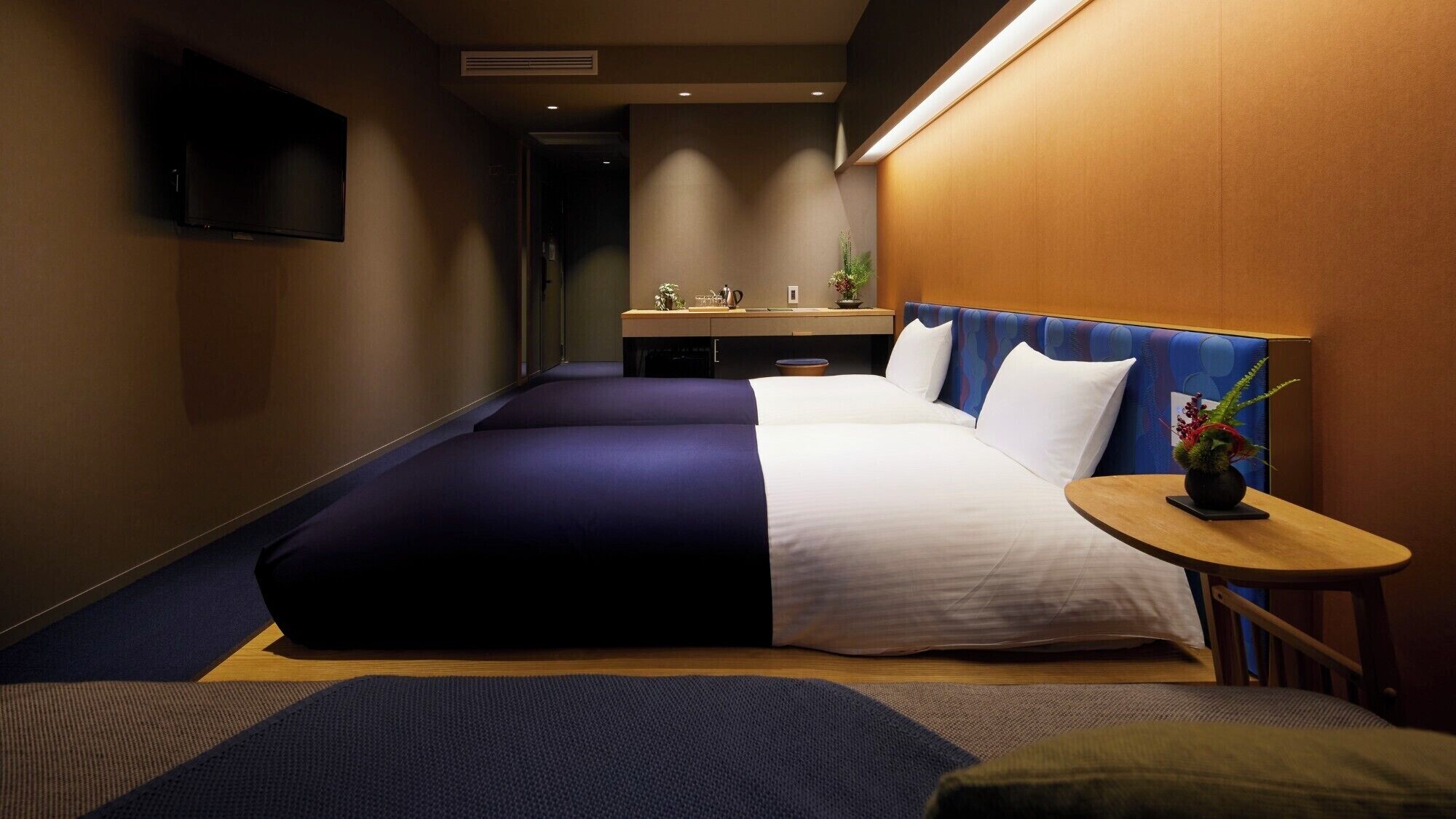 A hotel room with a large bed, a small desk, a TV mounted on the wall, and a small table with a plant.