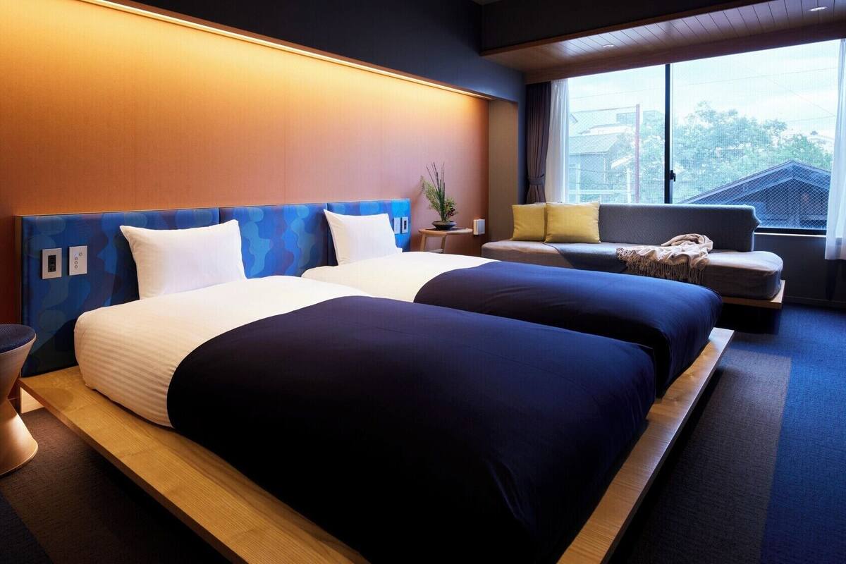 Hotelkamer met twee bedden, een bank, een klein tafeltje met een plant en een raam met uitzicht op gebouwen.