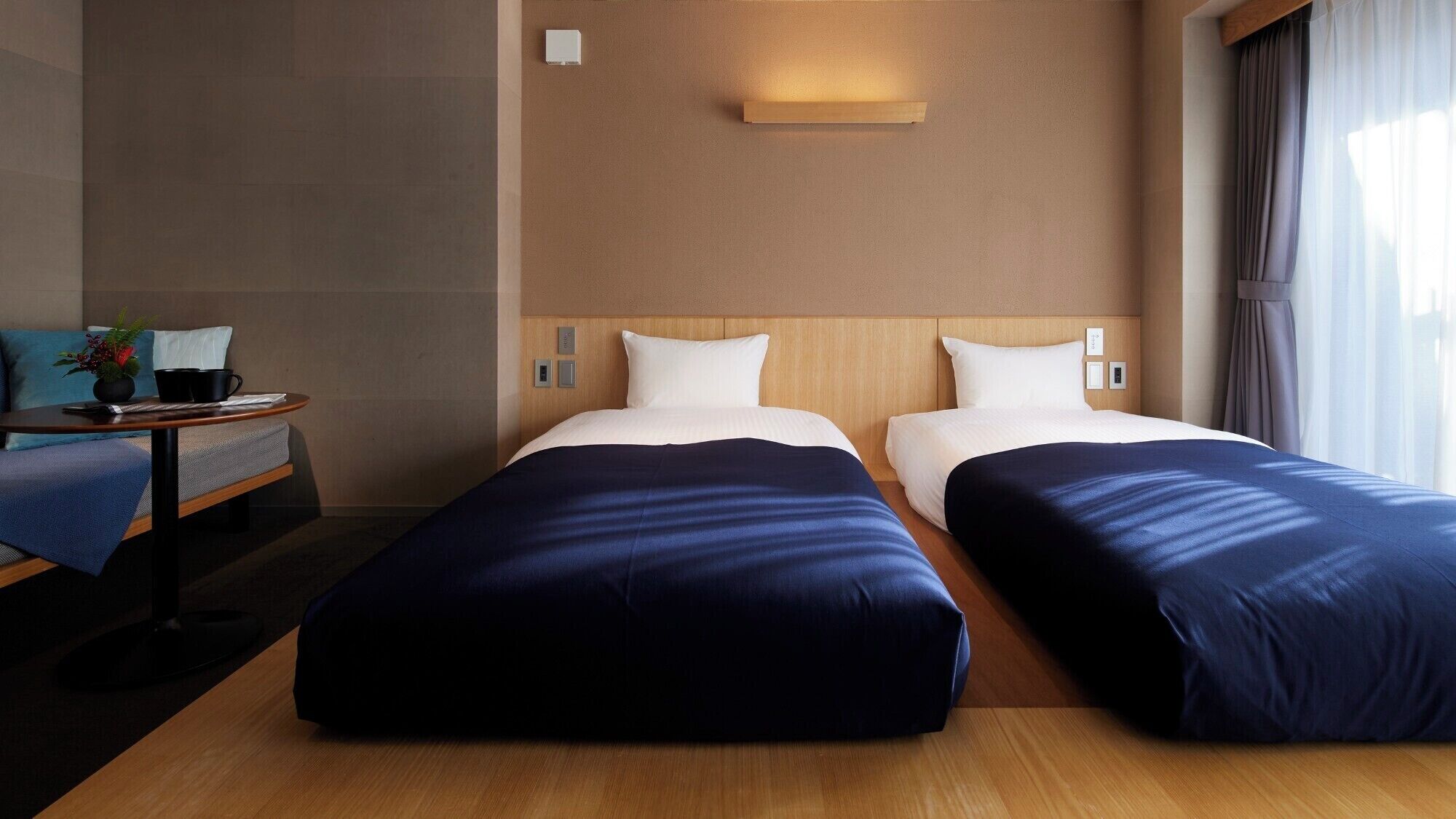 A hotel room with two beds, a small table with a plant, and a view of the outside through a window.