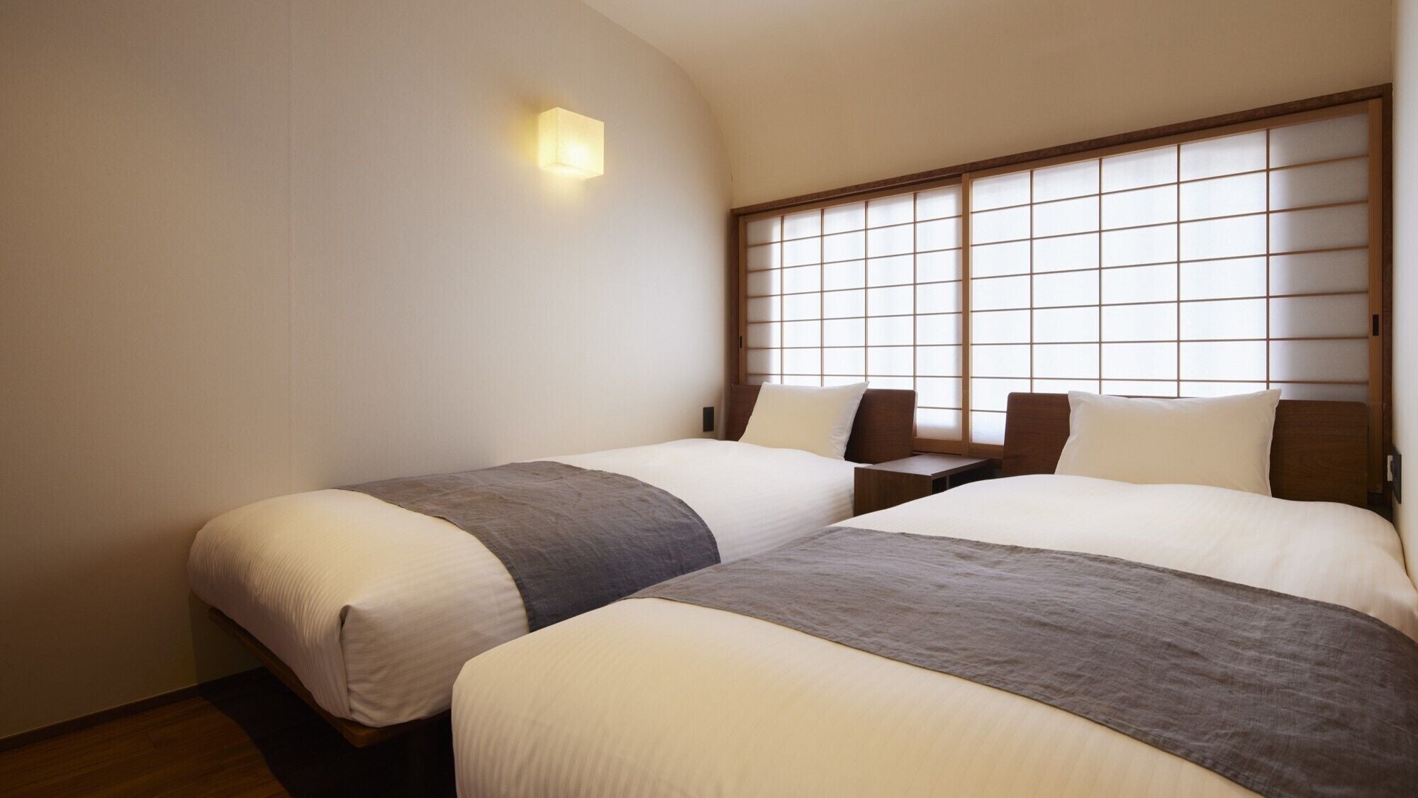 A hotel room with two beds, a nightstand, a wall-mounted light, and a shoji screen.