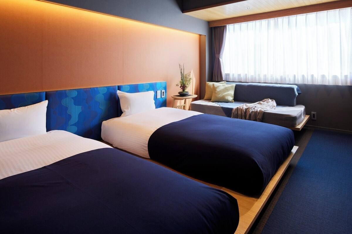 Een hotelkamer met twee bedden, een bank, een kleine tafel met een plant en een raam met gordijnen.