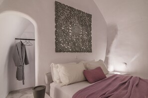 A bedroom with a bed, a hanging robe, and a decorative wall panel. - Yposkafon Concept (Santorini)