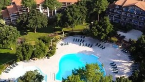 An aerial view of a resort with a central swimming pool surrounded by lounge chairs, a building with a red roof, and lush greenery. - Sunny terrace overlooking golf course and forest.★ (Moliets et Maâ)