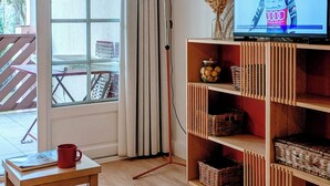 A living room with a television displaying a marathon runner, a wooden table with a red mug, and a large window with curtains. - Sunny terrace overlooking golf course and forest.★ (Moliets et Maâ)
