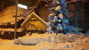 A snow-covered outdoor nativity scene with a star above the manger, surrounded by snow and a Christmas tree with lights.