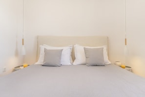Un lit bien fait, recouvert d’une couverture grise, avec deux oreillers blancs et deux oreillers gris. Il y a deux tables de chevet avec des lampes, et un verre de jus d’orange est posé sur l’une d’elles.
